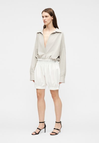 Γυναίκα που φοράει άνετο μπεζ μακρυμάνικο τοπ με βαθύ ντεκολτέ, λευκά σορτς με ελαστική μέση και μαύρα ψηλοτάκουνα σανδάλια με λουράκια, όρθια στάση.