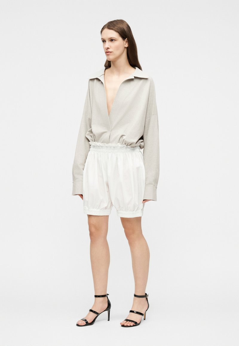 Femme portant un haut ample beige à manches longues et décolleté plongeant, un short blanc à taille élastique, et des sandales à talons hauts noires avec des lanières, debout.