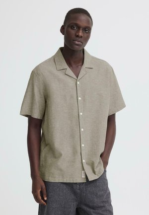 Young man with dark skin wearing a beige short-sleeve button-up shirt and dark gray pants, standing with one hand in pocket.