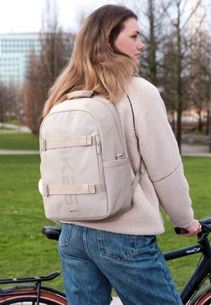 Young woman in beige fleece jacket and jeans wearing beige backpack, standing beside a black bicycle in a park with trees and buildings in background.