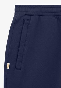 Sezione di pantaloni della tuta blu navy con elastico in vita, tasca verticale laterale e piccolo tag beige del marchio sulla cucitura sinistra.