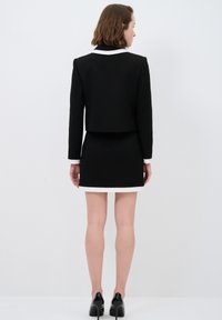 Svart kort blazer med vit krage, ihopparad med en svart minikjol med vit nederkant. Modellen har på sig svarta klackskor.