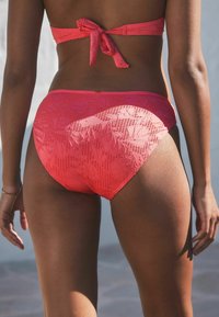 Korálkový bikini s květinovým krajkovým vzorem, s páskem zaväzovaným na chrbte a nízko vyrezanými nohavičkami. Tkanina je hladká a vzorovaná.