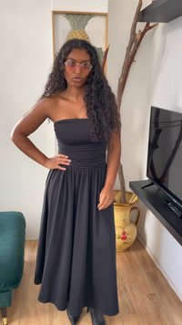 Melnā bezspoles maksi kleita, kas izgatavota no gludas auduma, ar sakrautu jostasvietu un plūstošu svārku. Vilkta kopā ar melniem potīšu zābakiem un apaļām rozā saulesbrillēm.