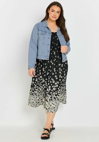 Giacca di jeans sopra un vestito floreale nero con margherite bianche, lunghezza a metà polpaccio. Il vestito presenta un corpetto arricciato e un tessuto fluido. Sandali neri.