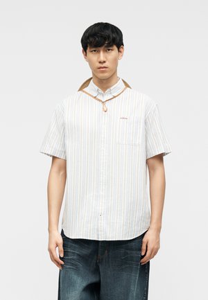 Vīrietis, kurš valkā īsām piedurknēm tumši svītrains krekls ar pogām un vaļīgas tumšas džinsas, stāv pret vienkāršu gaišu fonu.