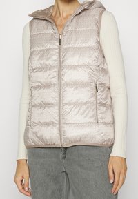 Chaleco acolchado de color rosa claro con capucha, presenta un patrón sutil y cierre delantero con cremallera. Dos bolsillos laterales y detalles acanalados en las mangas.