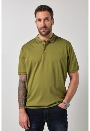 Mann mit Bart, der ein olivgrünes Poloshirt, dunkle Jeans, eine Armbanduhr und einen Tattoo-Ärmel am linken Arm trägt, steht vor einem schlichten Hintergrund.
