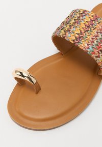 Sandale plate tissée avec une sangle multicolore, une semelle intérieure lisse couleur caramel et un anneau de doigt en métal doré.