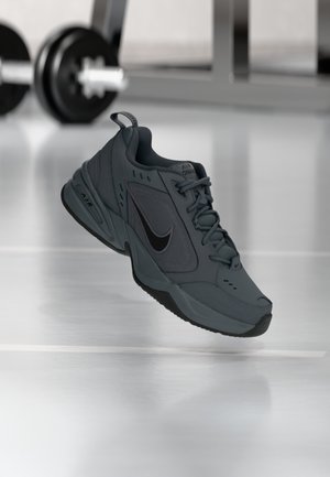 Baskets Nike Air Monarch gris foncé suspendues en l'air au-dessus d'un sol de gym lisse avec du matériel de musculation flou en arrière-plan.