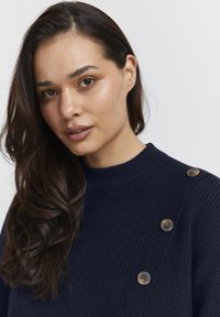 Námornícky rebrovaný sveter s vysokým golierom a bočnými gombíkmi. Látka má jemnú textúru a hladké, okrúhle gombíky.