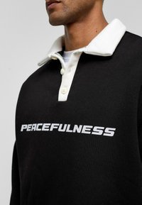 Zwarte sweatshirt met een witte kraag, met het woord "PEACEFULNESS" in vet, reflecterende letters op de voorkant. Zachte textuur.