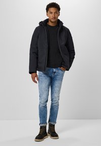 Black padded jacket with a hood, worn over a dark sweater, paired with light blue jeans and black lace-up boots. Simple design, visible stitching.