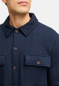 Marineblaue gesteppte Jacke mit einem Zickzackmuster, ausgestattet mit einem abgerundeten Kragen, Knopfverschluss und zwei Brusttaschen mit braunen Knöpfen.