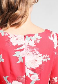 Robe florale rouge en tissu texturé, ornée de grands motifs de pivoines roses et blanches, avec une fermeture éclair au dos au niveau du col.