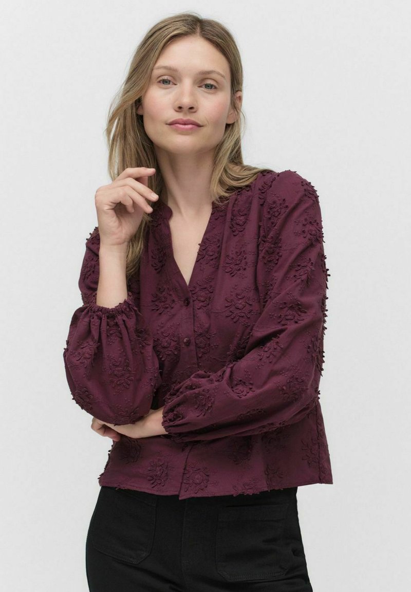 Bordeaux blouse met geborduurde bloemenstructuur, knoopsluiting aan de voorkant, lange, gezwollen mouwen en een relaxte pasvorm. Zwarte broek is ook zichtbaar.