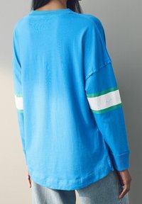 Vaaleansininen pitkähihainen paita, jossa vihreä- ja valkoraidalliset yksityiskohdat hihoissa. Pehmeä kangas, rento istuvuus, pyöreä kaula-aukko ja sivuhalkiot.