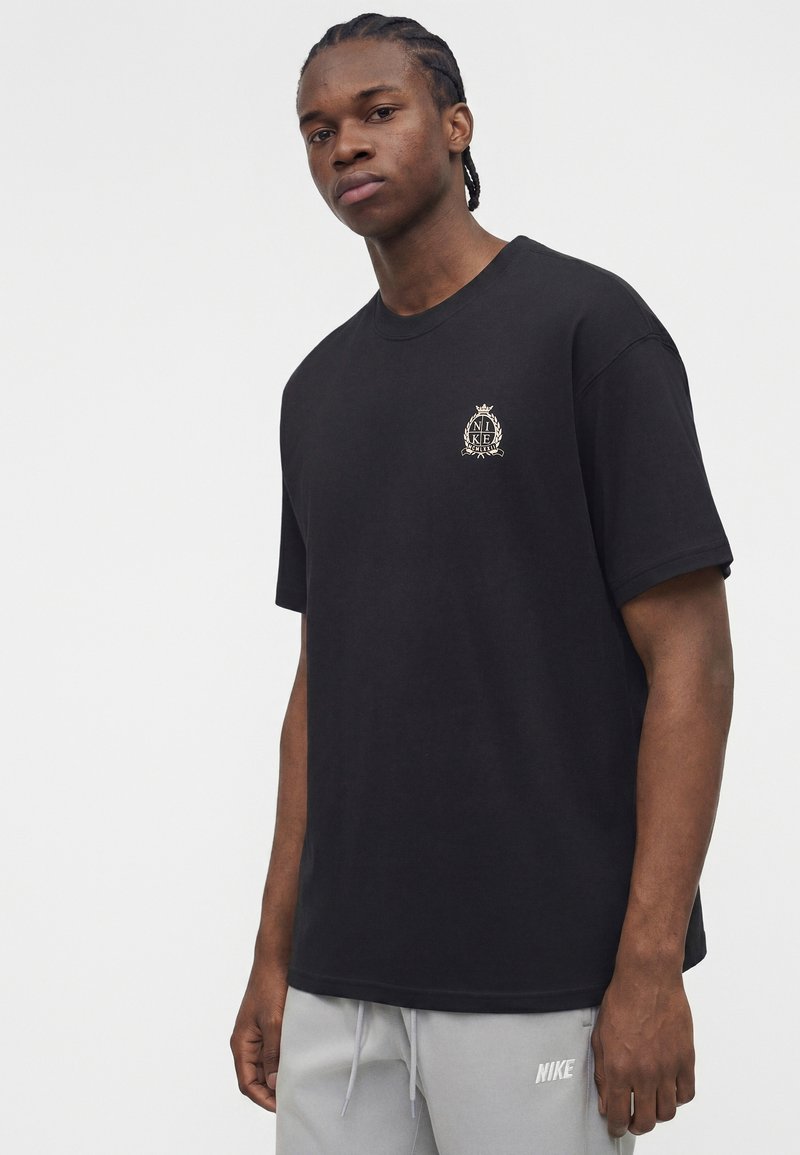 Giovane uomo con capelli intrecciati che indossa una t-shirt nera Nike con logo sul petto e pantaloni della tuta Nike grigio chiaro su sfondo uniforme.
