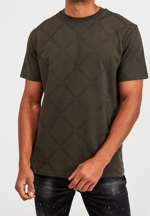 Homme portant un t-shirt à manches courtes vert olive foncé avec un motif en losanges subtil et du texte, associé à un jean noir avec des éclaboussures de peinture blanche.