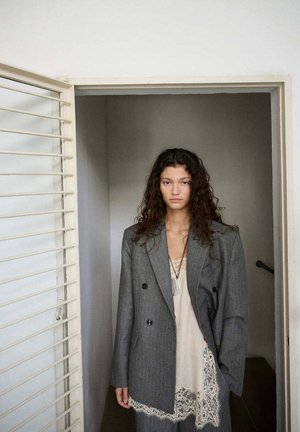 Femme aux longs cheveux bouclés portant un blazer gris oversize sur une robe en dentelle crème, debout dans un cadre de porte avec une expression neutre.