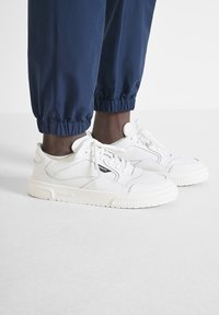 Sneakers atletici bianchi con un design testurizzato, chiusura con lacci e una suola di gomma spessa, abbinati a pantaloni jogger blu navy.