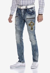 Hellblaue Jeans aus Denim mit dekorativen Nähten, Destroyed-Details und einem bestickten Kreuz-Design. Kombiniert mit einem weißen Nietengürtel.