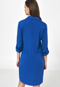 Niebieska sukienka koszulowa wykonana z gładkiego materiału, z kołnierzykiem, rękawami o długości trzech ćwierć i zaokrąglonym dołem z bocznymi guzikami.