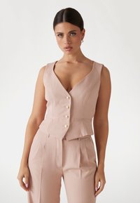Blush-pink ärmellose Weste mit V-Ausschnitt, versehen mit fünf Perlenknöpfen und taillierter Passform, kombiniert mit passenden Hosen. Glatte Stofftextur.