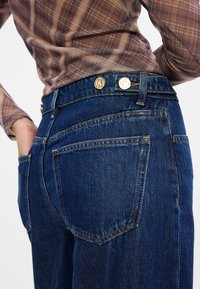 Jeans de denim azul escuro com uma cintura alta, botões dourados e bolsos traseiros clássicos. O tecido tem uma textura subtil e detalhes de costura visíveis.