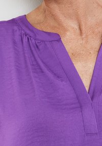 Lila blus med V-ringning, veckad detaljer vid axeln och slät, något glänsande tyg.