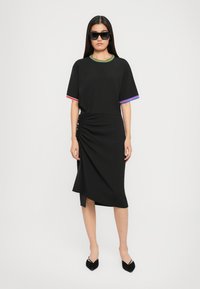 Kvinna som står i en svart rynkad klänning med flerfärgad kant vid ärmarna och halsringningen, iförd svarta spetsiga slingback-skor och solglasögon.