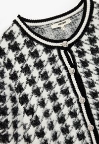 Svart och vit houndstooth kofta med en fluffig textur, svarta kanter och tre dekorativa knappar fram.
