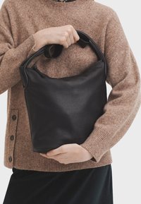 Sac en cuir noir avec une texture douce et des poignées arrondies, tenu par une personne portant un pull marron, présentant un design sans couture.