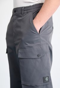 Gray cargo pants with an elastic waistband, side pockets, and a flap pocket featuring a black button and brand patch. Smooth, durable material.