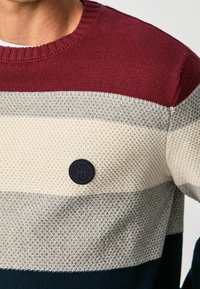 Gebreide trui met horizontale strepen in bordeauxrood, crème, grijs en marineblauw. Heeft een gestructureerd patroon en een zwarte ronde logo-patch.