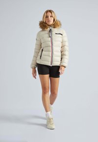 Pufferjack in lichtbeige met een nepbontkap, ritszakken en afwisselende verticale strepen aan de voorkant. Draagt zwarte shorts.