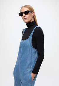 Lys blå denim-dress med løs passform, rund hals og sidelommer, brukt over en tettsittende svart turtleneck. Sorte solbriller.