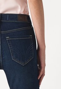 Jeans de mezclilla azul oscuro con un diseño ajustado, que cuentan con dos bolsillos traseros y detalles de costura en contraste. Etiqueta de cuero marrón en la cinturilla visible.