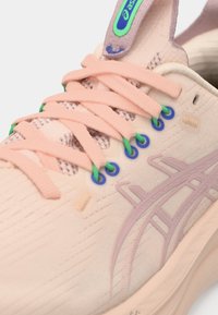 Chaussure de sport rose clair avec surface texturée, présentant des accents violets et des œillets verts et bleus. Les lacets sont roses et bien serrés.