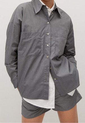 Woman wearing oversized gray button-up shirt with large front pockets over a white shirt, paired with matching gray shorts.