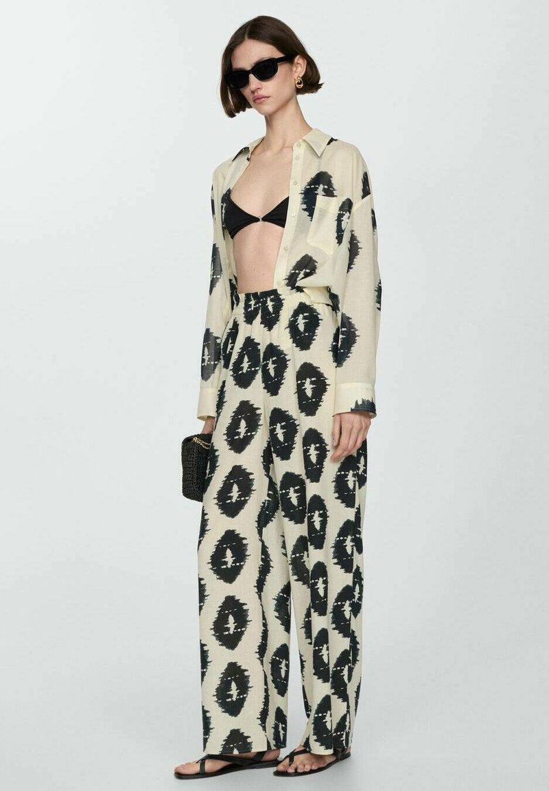 Leichtes, cremfarbenes Hemd mit großem schwarzem Ikat-Muster, kombiniert mit lockeren schwarzen und cremfarbenen Hosen. Ein schwarzes Bikini-Oberteil, Sonnenbrille und Tasche runden den Look ab.