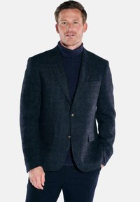 Marineblauer Wollblazer mit strukturiertem Stoff, einreihiges Design, zwei Fronttaschen und braunen Knöpfen. Trägt man über einem dunklen Rollkragenpullover.