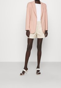 Blazer ajustado de color rosa claro con solapas en pico, top blanco, pantalones cortos beige y sandalias de tacón blancas, con una textura suave y un diseño limpio.