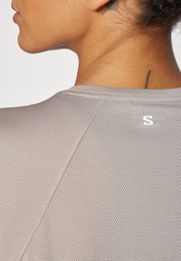 Camisa atlética de malla gris con cuello redondo y un logo sutil en la parte posterior. La tela tiene un patrón texturizado para la transpirabilidad.