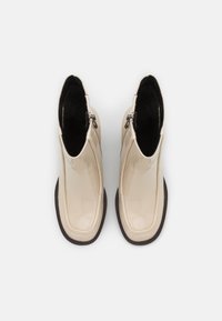 Stivali ankle in pelle verniciata beige con un design elegante, chiusura con zip e suola in gomma scura texturizzata. Finitura liscia con punta arrotondata.