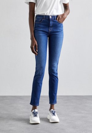 Personne portant un jean slim bleu foncé, un T-shirt blanc et des baskets blanches, debout sur un sol gris contre un mur clair uni.