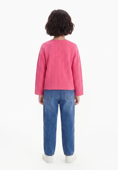 Enfant aux cheveux courts et bouclés, debout de dos, portant un haut rose à manches longues texturé, un jean bleu et des chaussures blanches sur fond blanc.