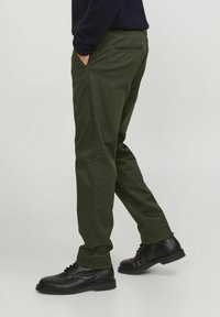Pantaloni de culoare verde măsliniu cu un croi slim, prevăzuți cu buzunare laterale și un buzunar cu fermoar la spate, asortați cu pantofi negri cu șireturi.