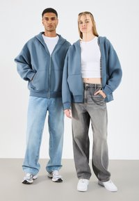 Blauwe hoodies met rits aan de voorkant, grote zakken en geribde manchetten, gecombineerd met losse, vervaagde denim jeans. Model draagt witte sneakers.