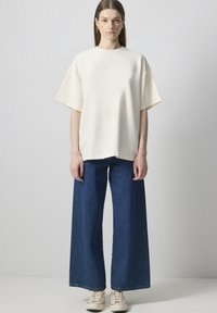 Helle beige topp med korte ærmer, parret med brede, blå denim jeans og lyse sneakers. Glat stofstruktur.